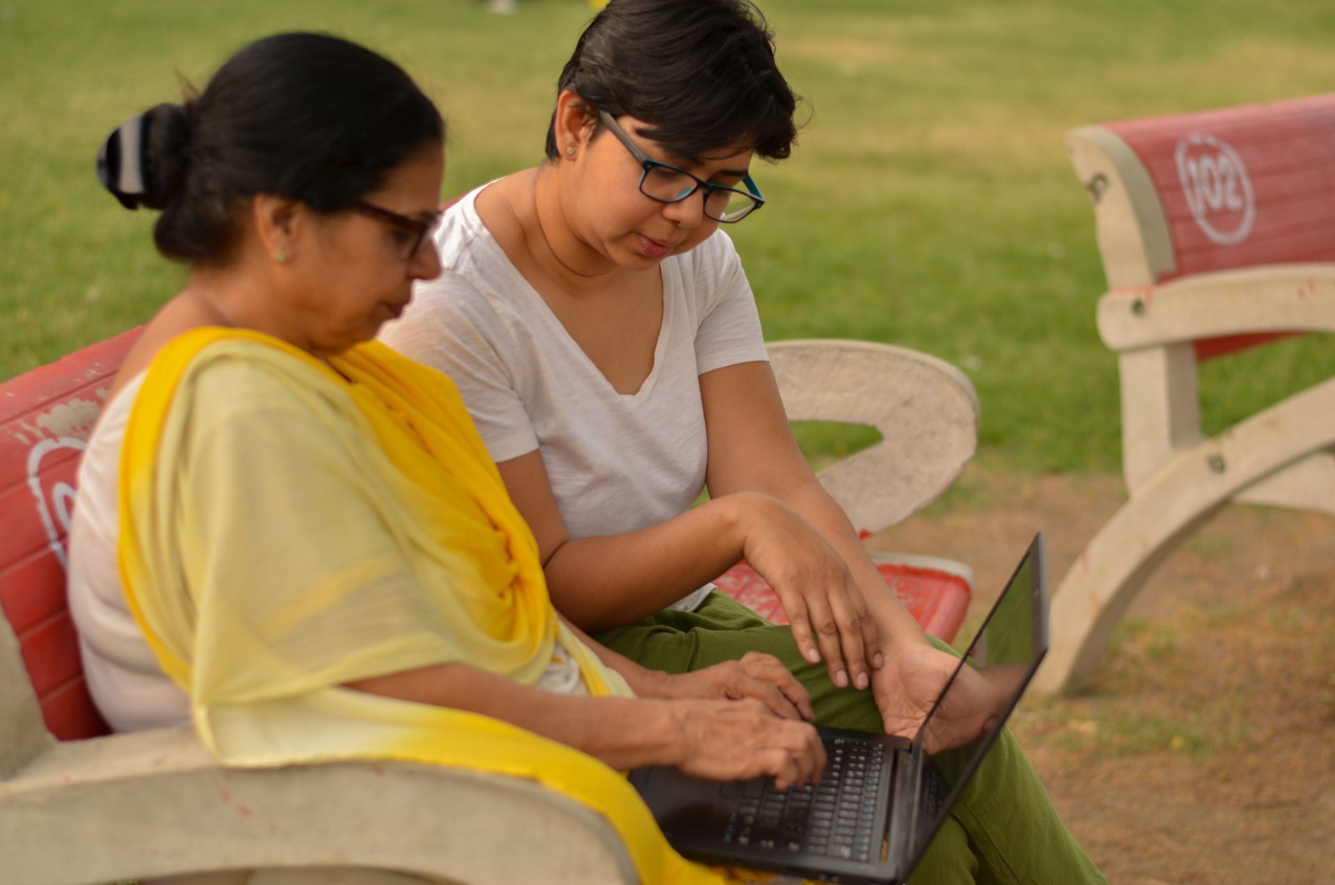 Side view of young Indian girl helping an old Indian woman on a laptop sitting on a red bench in a park in Delhi, India. Concept Digital literacy / Education